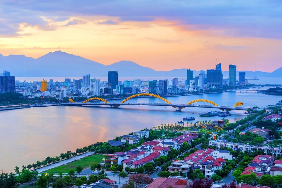 Golden Dragon Bridge illuminated at sunset over Han River with Danang city skyline and mountains in background