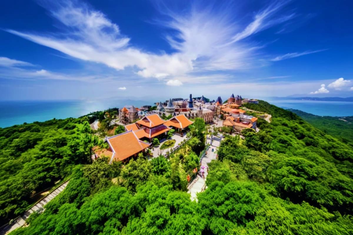 Aerial view of Ba Na Hills castle complex with orange-tiled roofs surrounded by dense green forest and coastal landscape in D