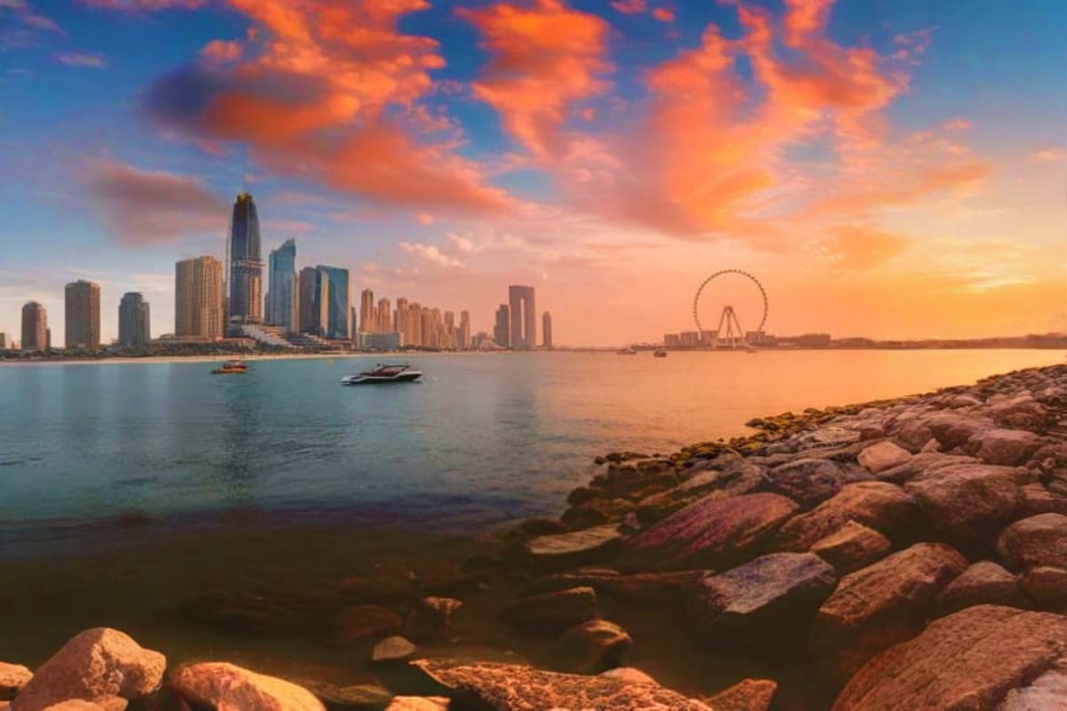 Dubai skyline at sunset with Burj Khalifa, marina boats, and Ferris wheel viewed from rocky shoreline