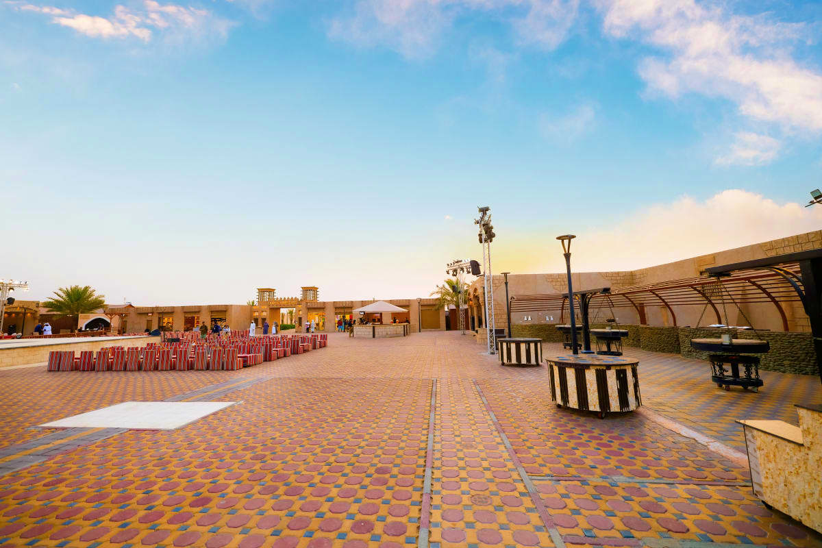 Dubai Desert Safari with Dune Bashing Traditional Arabic courtyard with beige buildings, palm trees, and patterned stone flooring under clear blue sky