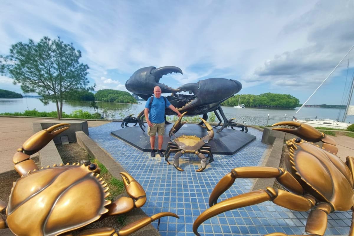 Enroute Krabi city tour to Phuket Man posing with large golden crab sculptures at waterfront promenade overlooking river and sailboats