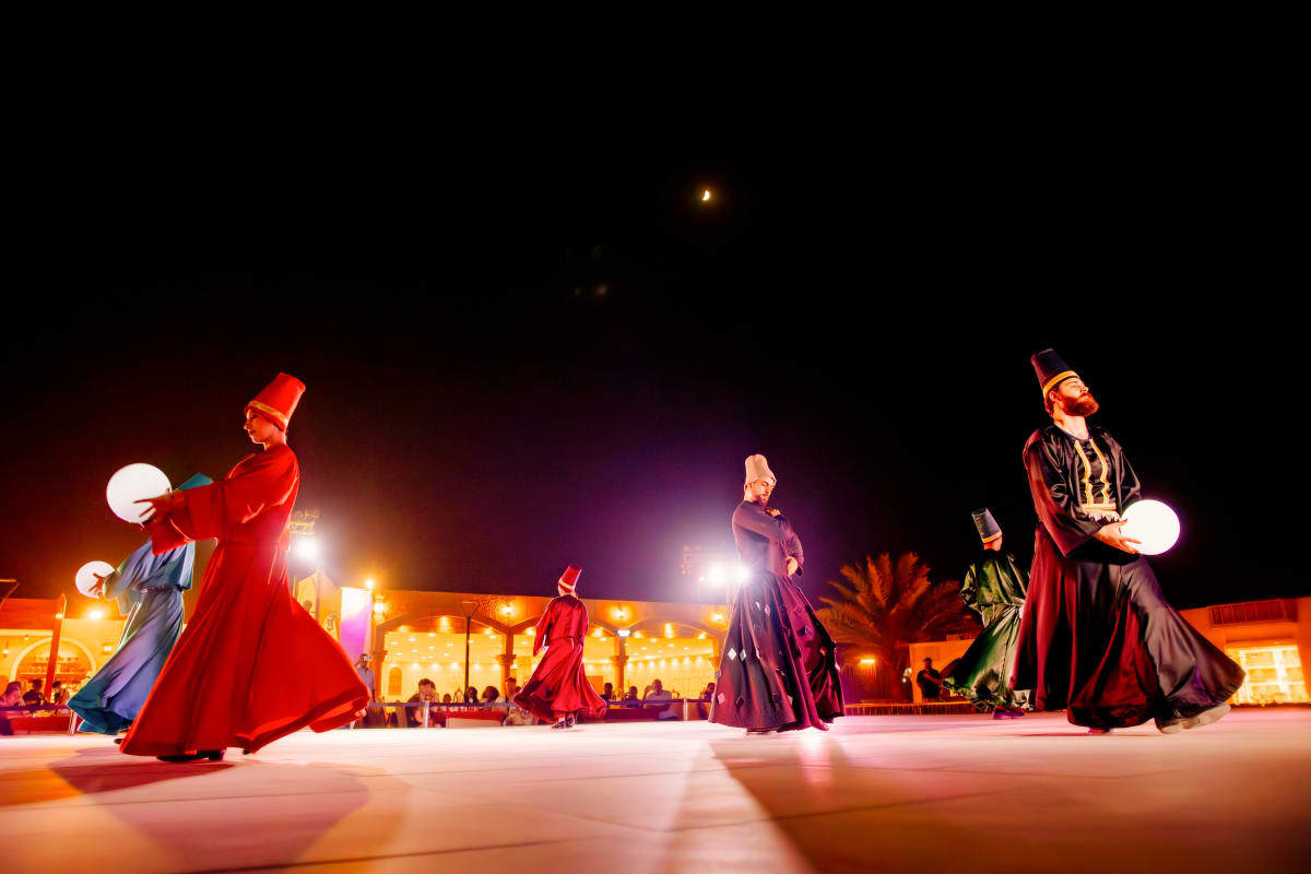Performers in traditional red and dark robes with illuminated globes on desert stage at night during evening entertainment sh