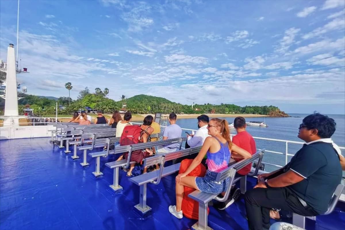 Passengers seated on an open-air ferry deck overlooking tropical coastline with palm trees and mountains under blue sky