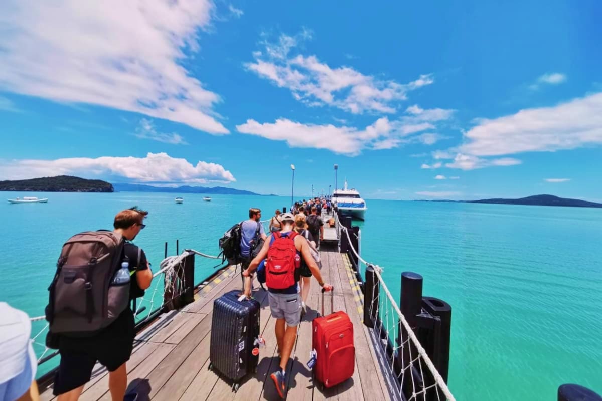 Tourists boarding a ferry at a wooden pier with turquoise water, mountains, and islands visible in the background under blue 