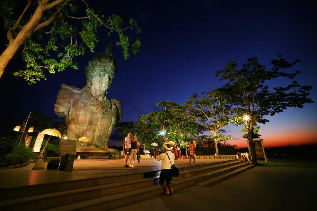 Illuminated stone statue of Garuda Wisnu Kencana at night with visitors walking on plaza surrounded by trees at dusk in Bali