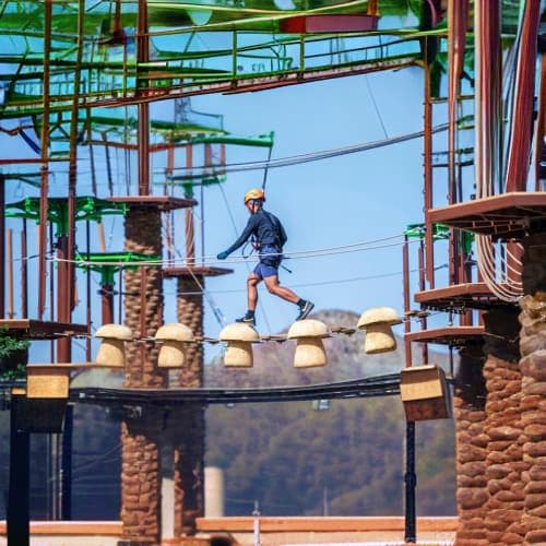 Person zip-lining over trampoline at Hatta adventure park with metal framework structures and palm trees visible