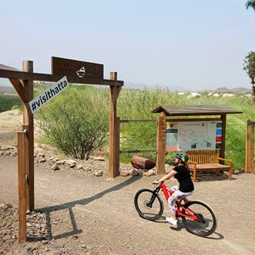 Cyclist on red bike at Wadi Hatta entrance sign with rest shelter and information board in desert mountain landscape