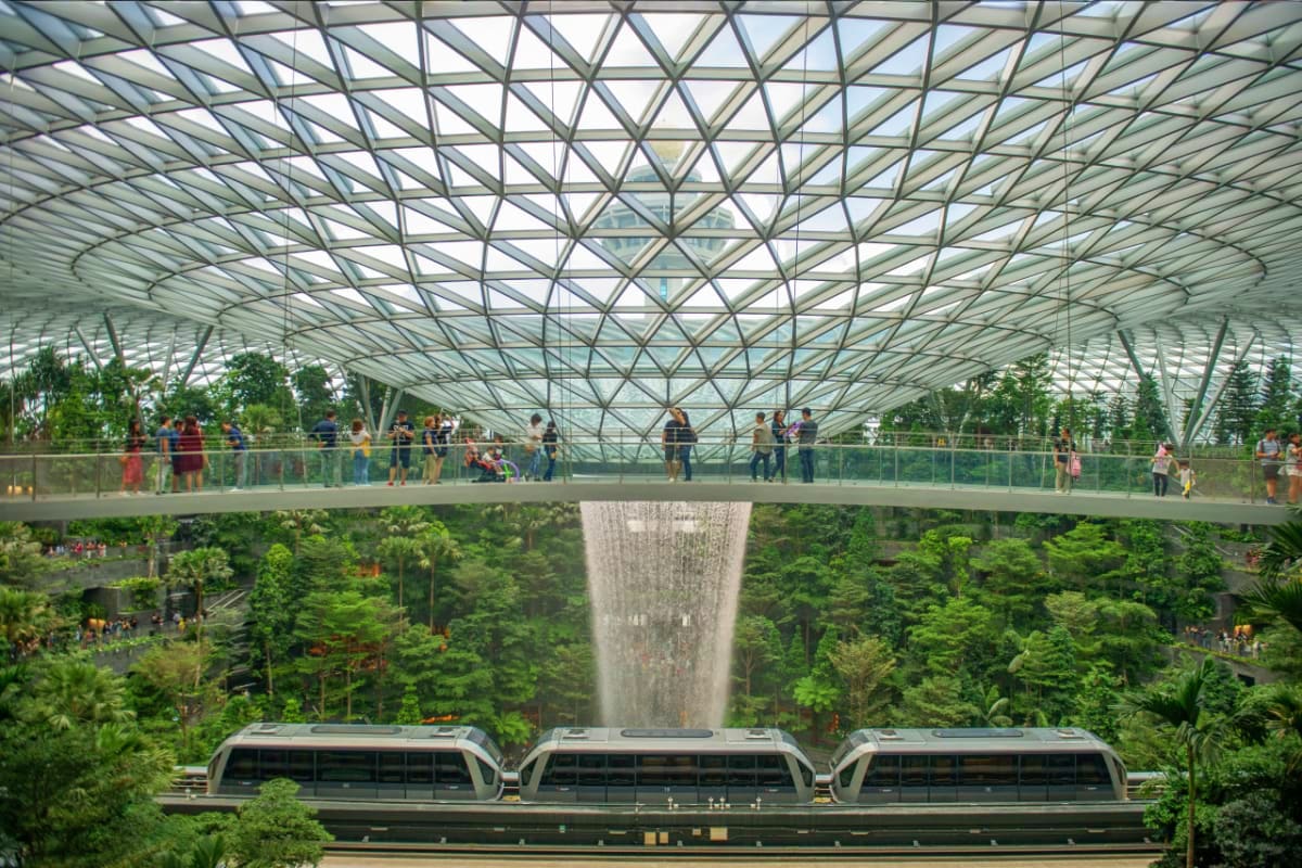 Indoor rainforest with indoor waterfall, lush green vegetation, glass ceiling, and train passing below at Jewel Changi Airpor