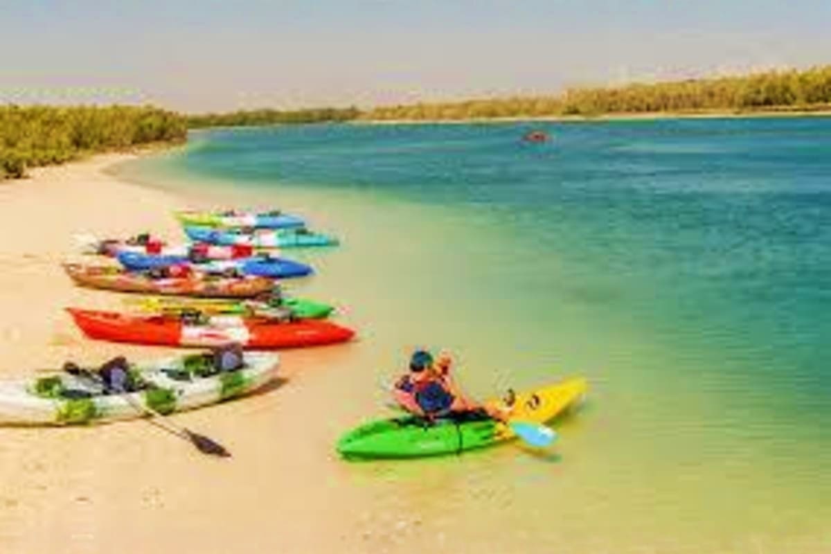 Person kayaking in turquoise water near sandy beach with colorful boats lined up on shore and mangrove trees in background