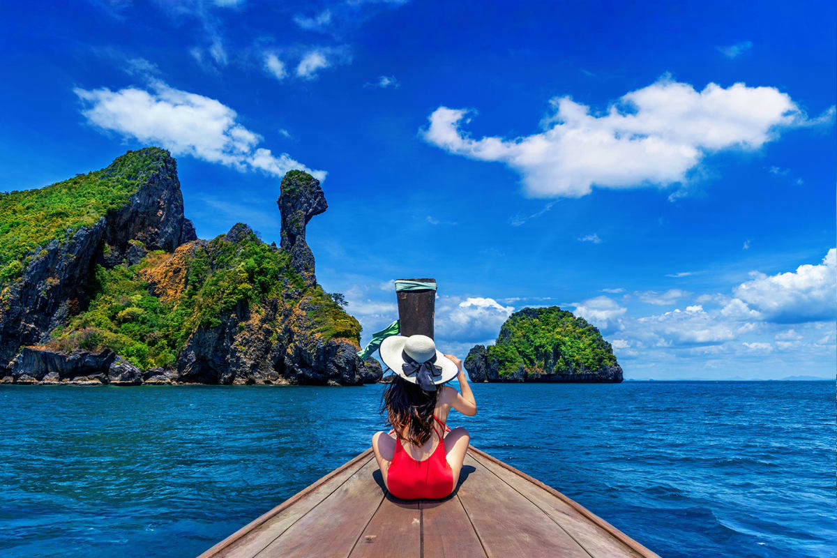 Krabi Island Tour from Phuket Woman in red swimsuit and white hat sitting on longboat bow facing limestone cliffs and turquoise water in Krabi, Thailand