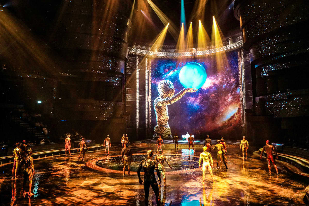 La Perle by Dragone Dubai Acrobatic performers in metallic gold bodysuits on wet stage floor with giant illuminated sphere and cosmic projection backdr