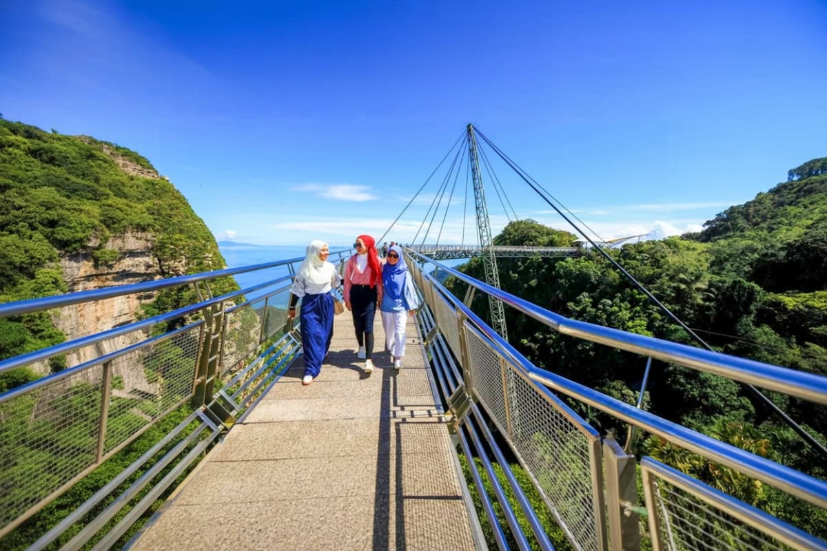 Langkawi Cable Car Three tourists walking on the Langkawi Sky Bridge suspended cable car walkway with blue railings, lush green cliffs and ocean