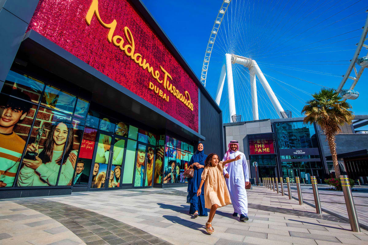 Family walking outside Madame Tussauds Dubai wax museum with red illuminated sign and Burj Jumeirah bridge visible in backgro