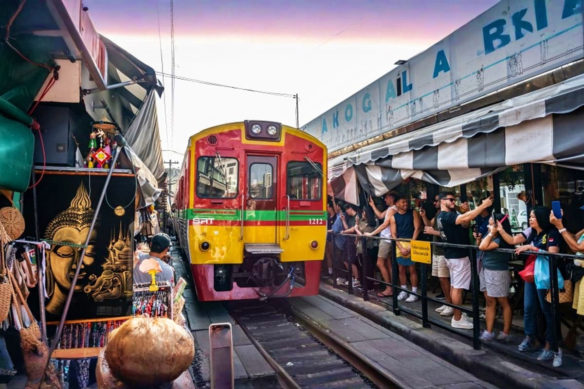 Red and yellow train numbered 1212 arriving at Maeklong Railway Market with vendors and shoppers along the tracks at sunset