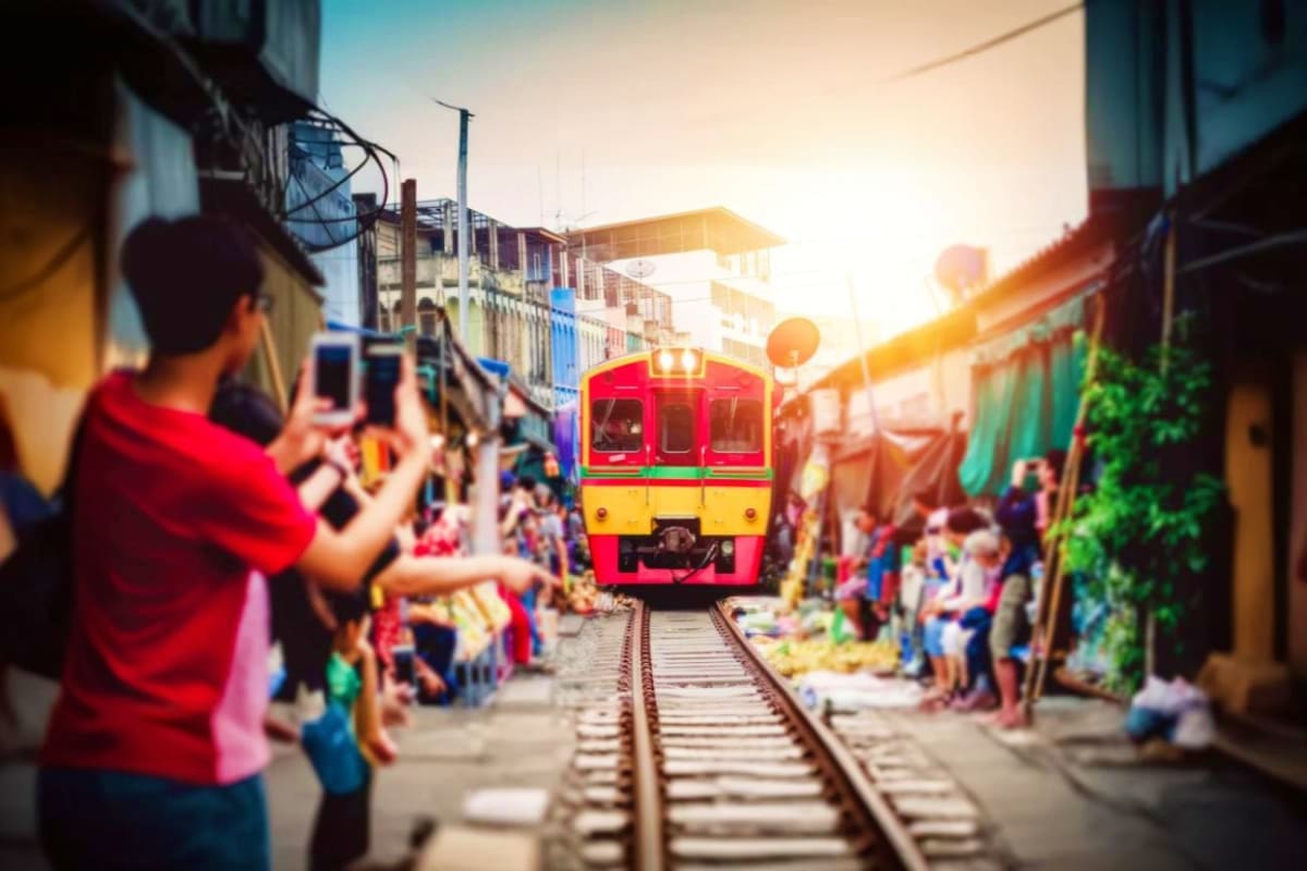 Red and yellow train approaching through crowded Maeklong Railway Market with tourists taking photos at sunset