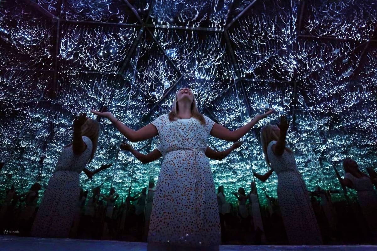 Woman with arms raised standing in illuminated room with glowing ceiling and mirrored figures at Museum of Illusions