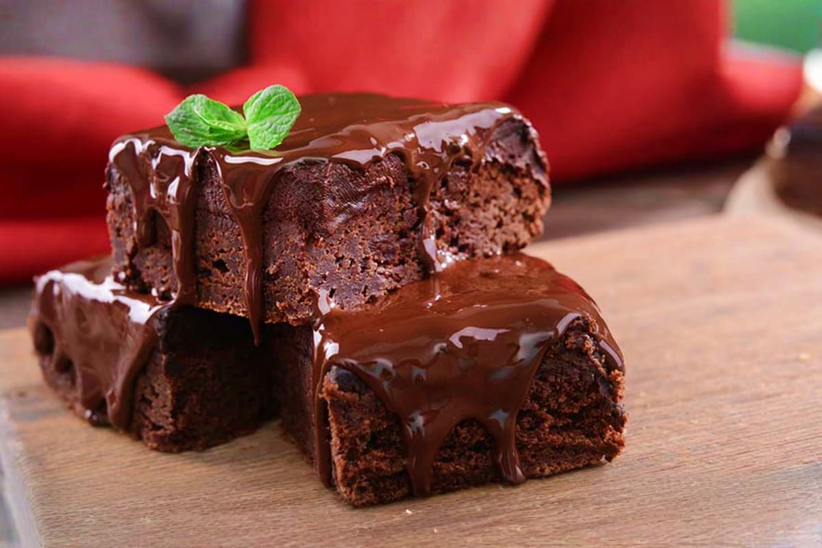 Rich chocolate cake slices with glossy ganache and fresh mint garnish on wooden surface with red cushions in background
