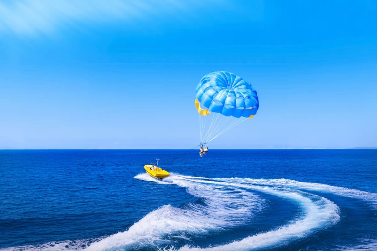 Person parasailing with blue and white parachute being towed by yellow speedboat off Cenang Beach with ocean waves