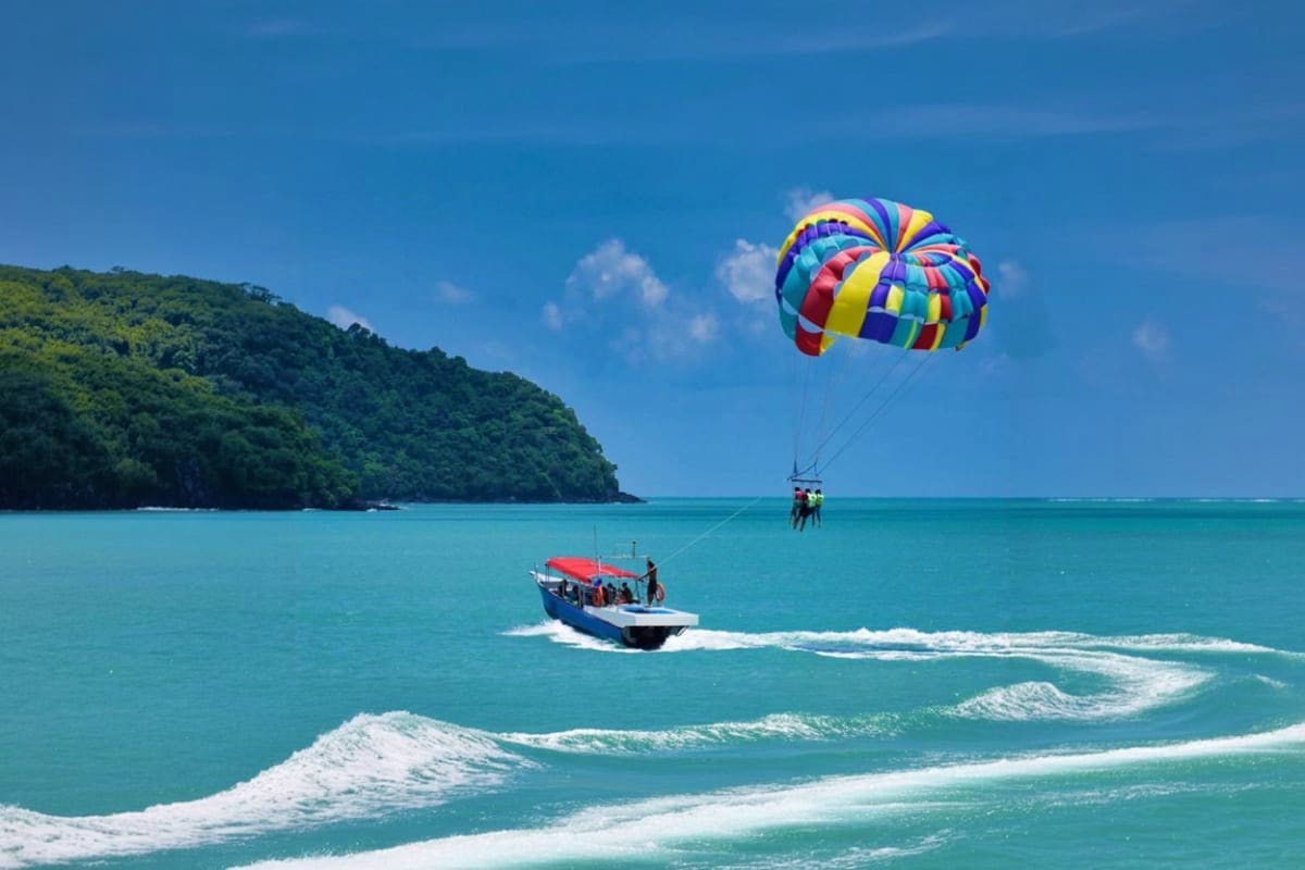 Colorful parasail with multiple people being towed behind a speedboat in turquoise water at Cenang Beach, with forested hills