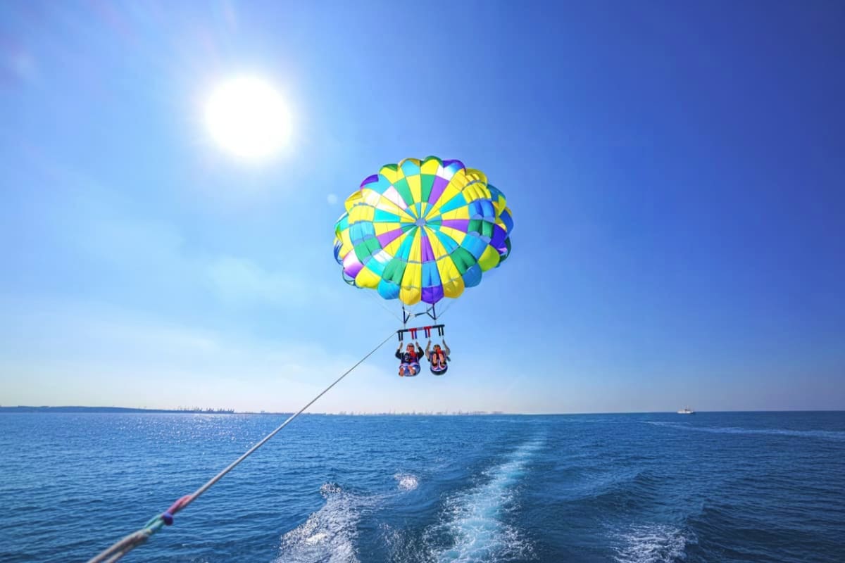 Colorful parasail with yellow, blue, and purple panels flying above the ocean with two passengers attached, towed from a boat
