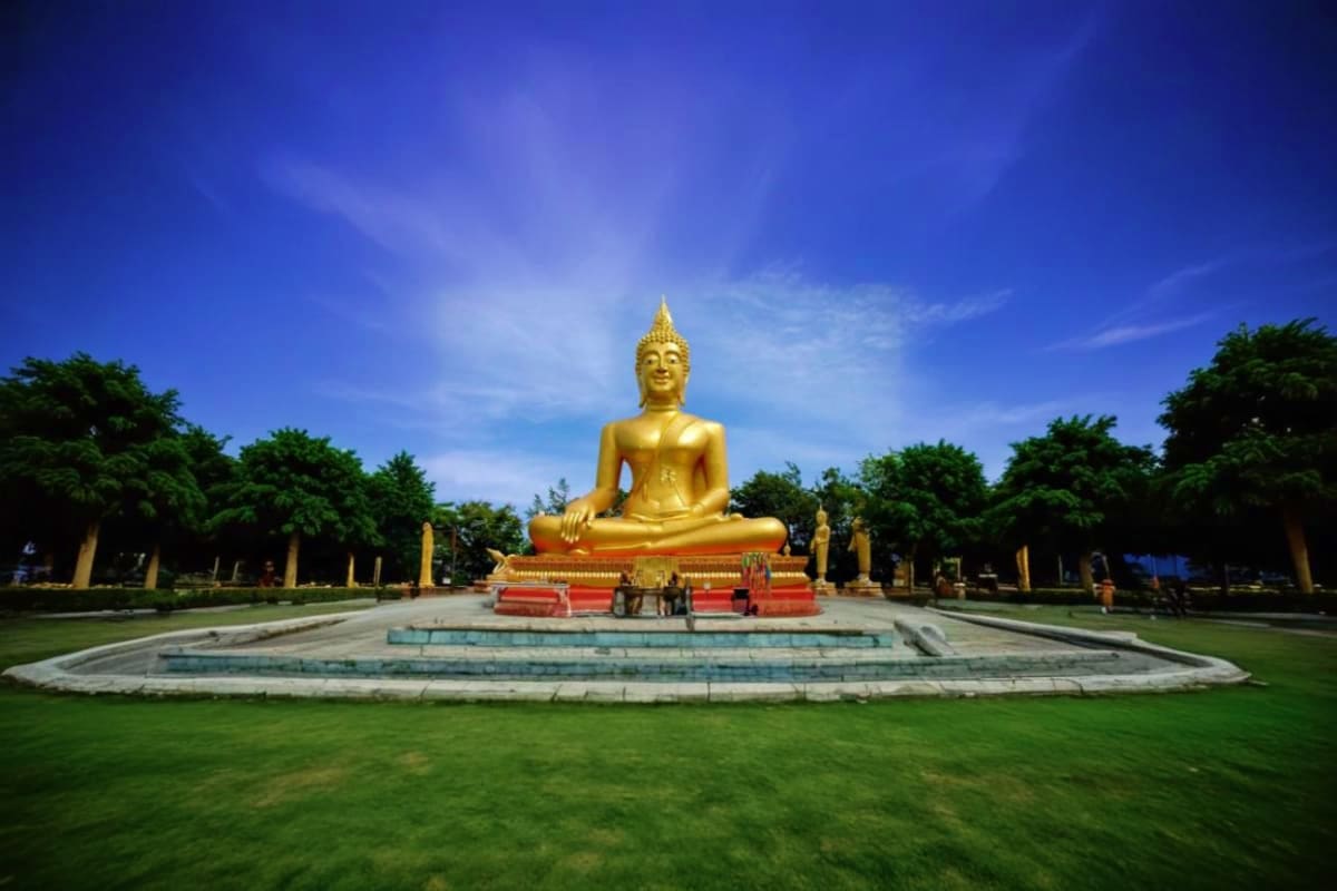 Large golden Buddha statue in seated meditation pose on red platform surrounded by green lawn and trees under blue sky in Pat