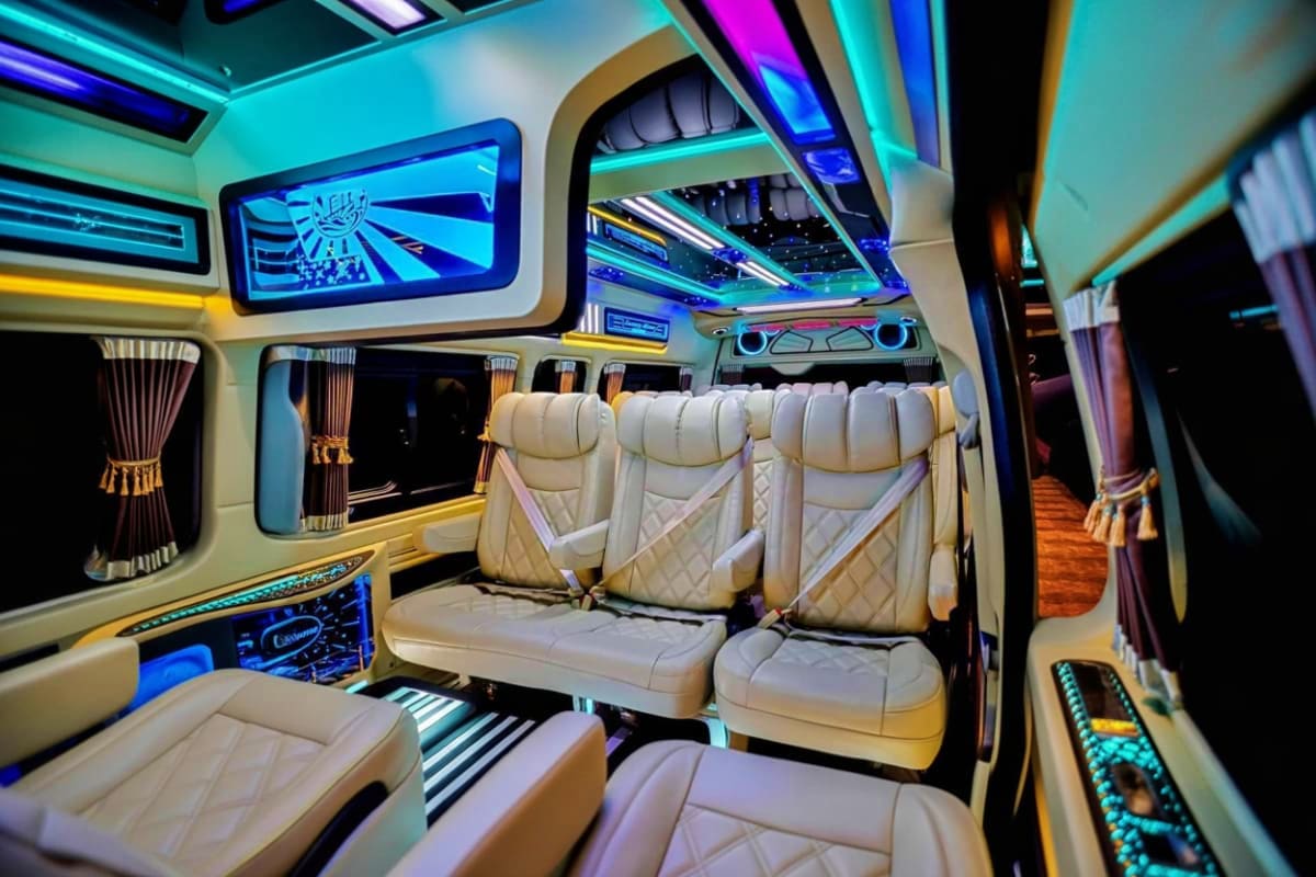 Luxury minibus interior with cream leather seats, cyan and blue neon lighting, digital displays, and gold trim accents