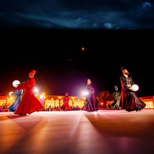 Performers in traditional red and colorful robes dancing with glowing lanterns at night in a desert setting with illuminated 