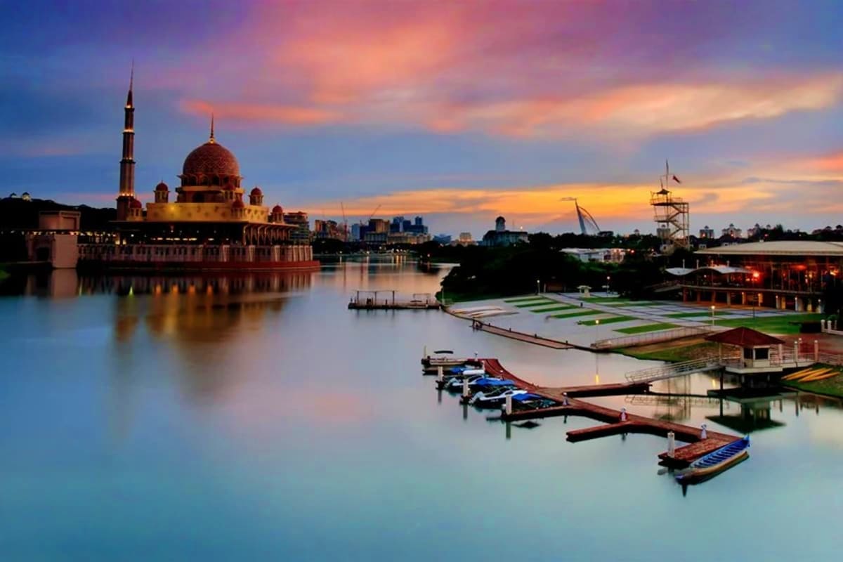 Putrajaya mosque with red dome at sunset reflected in calm water, traditional wooden boats docked in foreground, city skyline