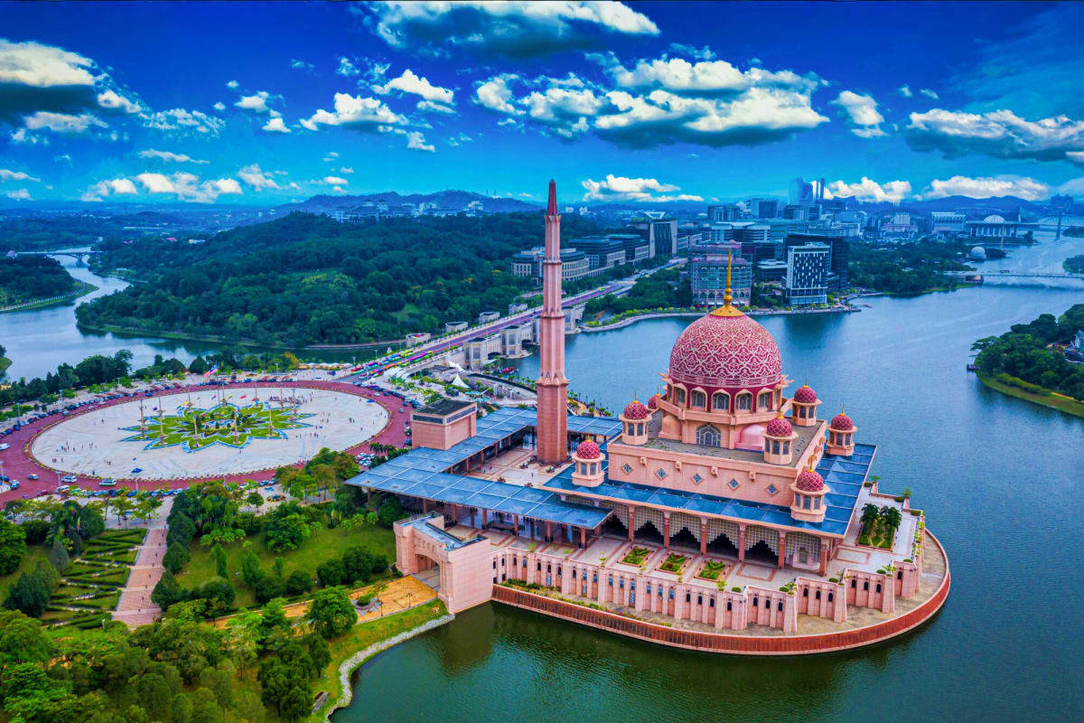 Aerial view of pink Putra Mosque with domed roof and minaret on island, surrounded by blue water, with city skyline and green