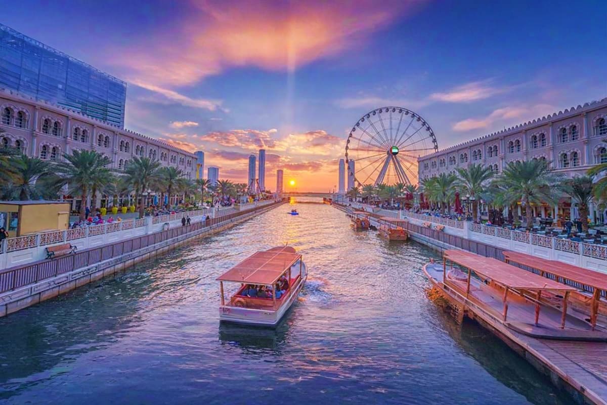 Sunset view of Sharjah waterfront with traditional abra boats, Ferris wheel, palm trees, and historic architecture along the 