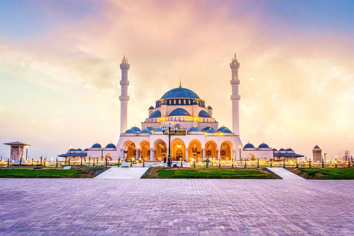 Grand mosque with blue domes and minarets illuminated at sunset in Sharjah, UAE