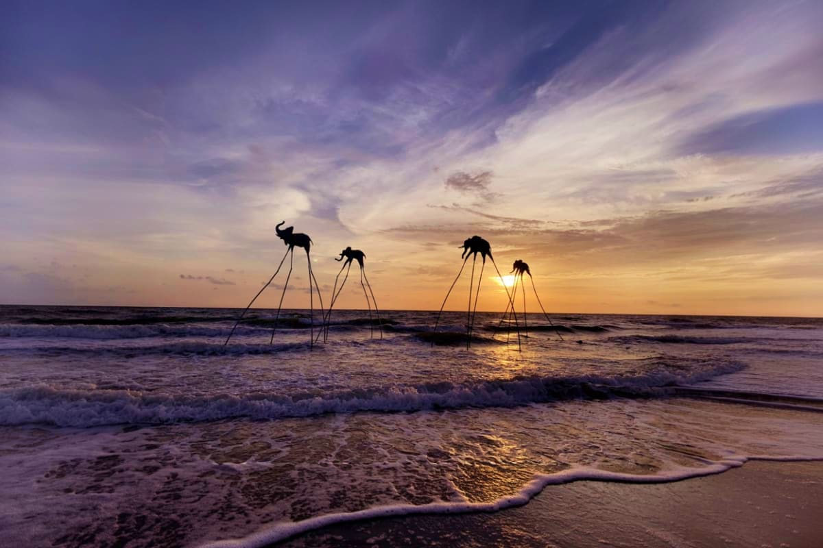Silhouetted sculptures of giraffes with long legs wading in ocean waves at sunset on beach