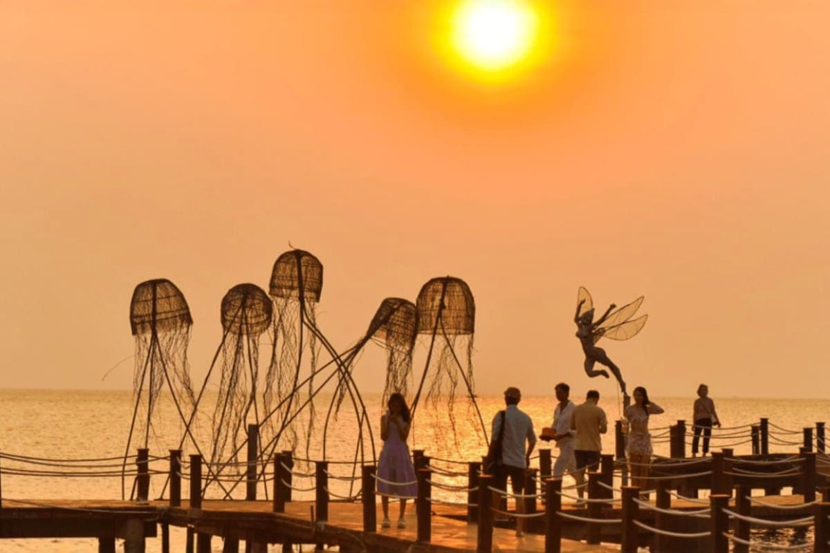 Silhouetted people on wooden pier at sunset with jellyfish sculptures and flying figure statue against golden sky and ocean