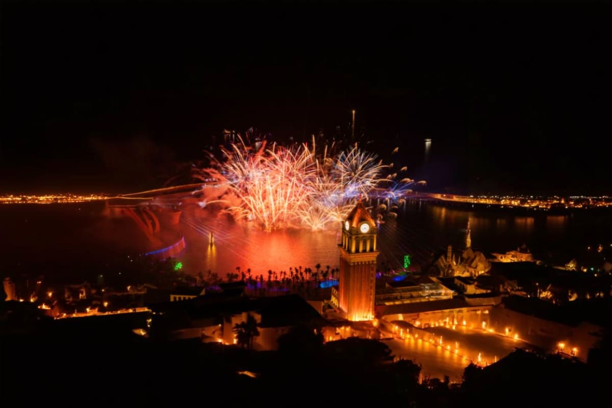 Fireworks display exploding over illuminated waterfront temples at night with crowds watching from shore during Symphony of t