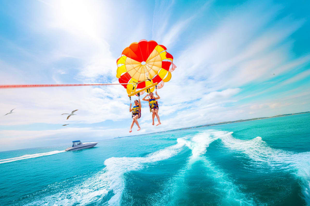 Tanjung Benoa Water Sports Two people parasailing on red and yellow parachute over turquoise ocean water with speedboat below at Tanjung Benoa beach