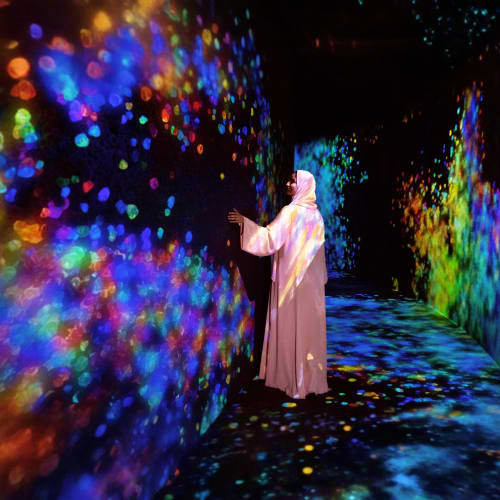 Person in white robe interacting with colorful digital light projections on walls in immersive art installation