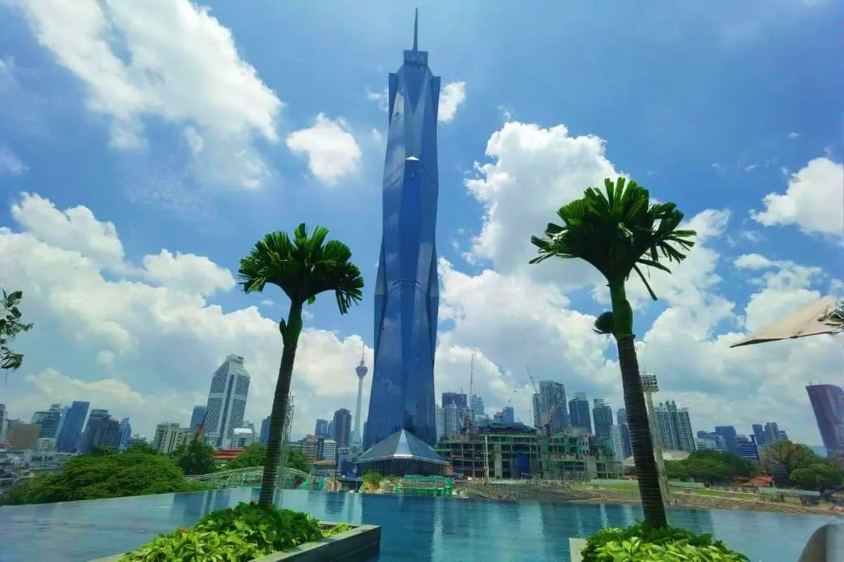 The Great Kuala Lumpur Iconic Attractions Tour Modern blue tower with sharp spire rising behind turquoise lagoon, flanked by palm trees, with Kuala Lumpur city skyline in b