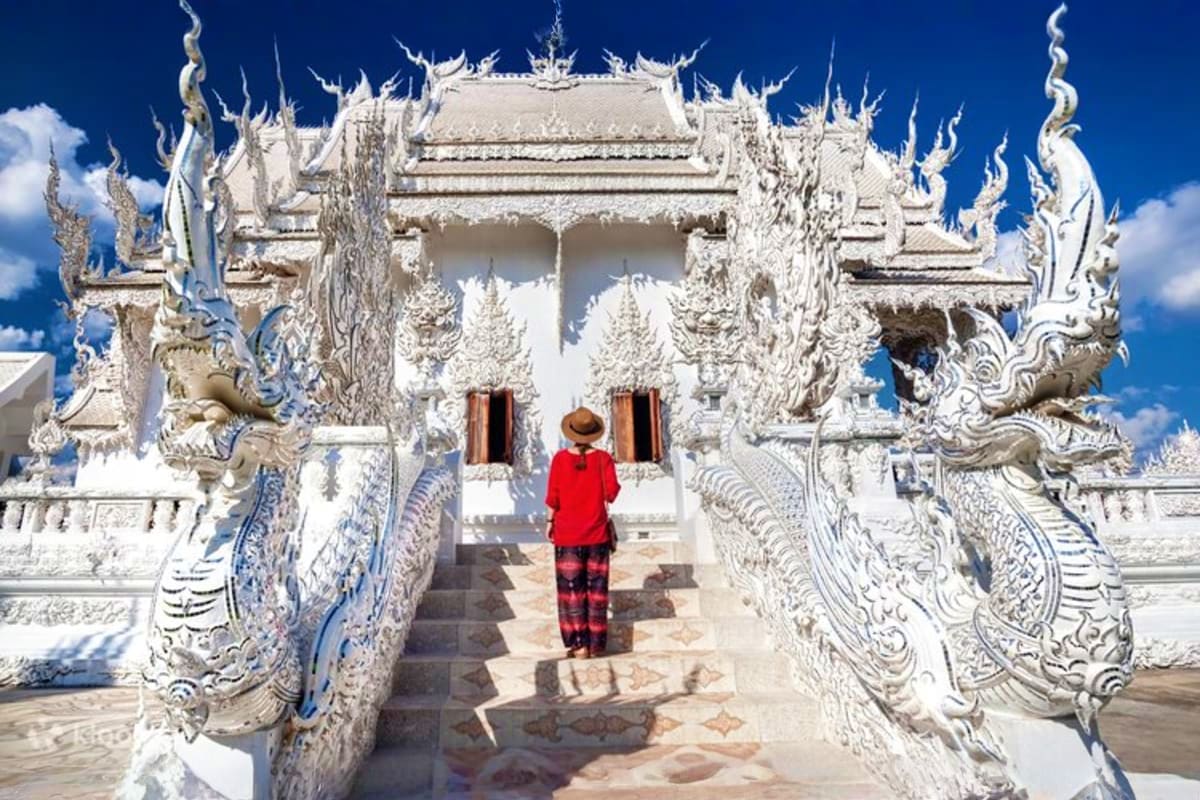 Tourist in red jacket walking through ornate white temple with carved dragon railings and traditional Thai architecture again
