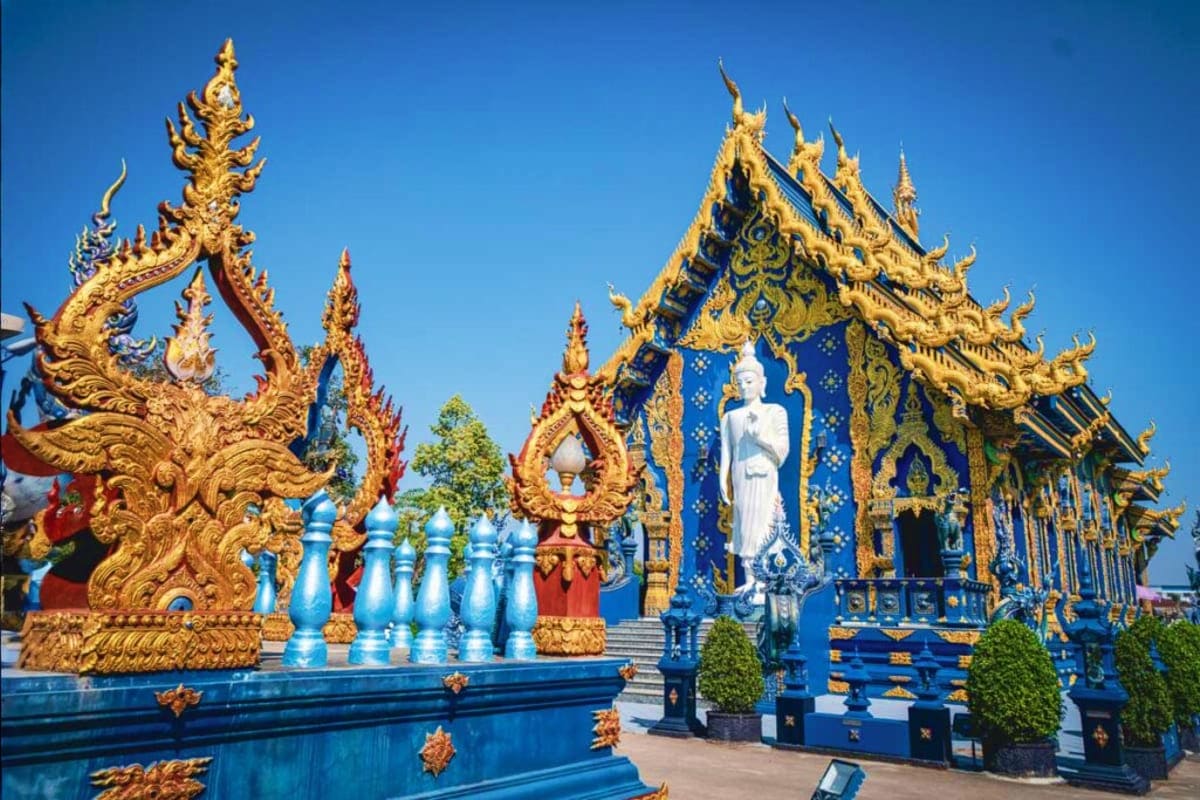 Ornate Thai Buddhist temple with golden roof spires, blue walls, and white Buddha statue under clear sky