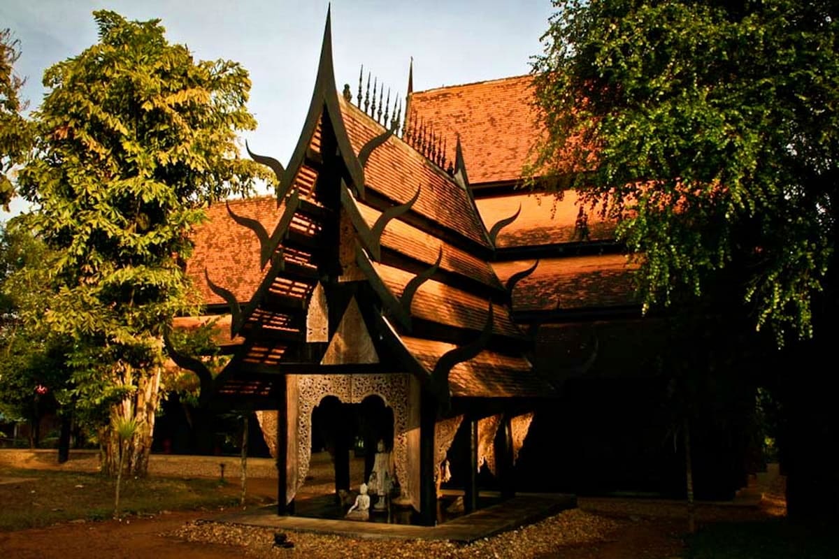 Traditional Thai temple with distinctive pointed black roof and ornate orange-tiled architecture surrounded by lush green fol