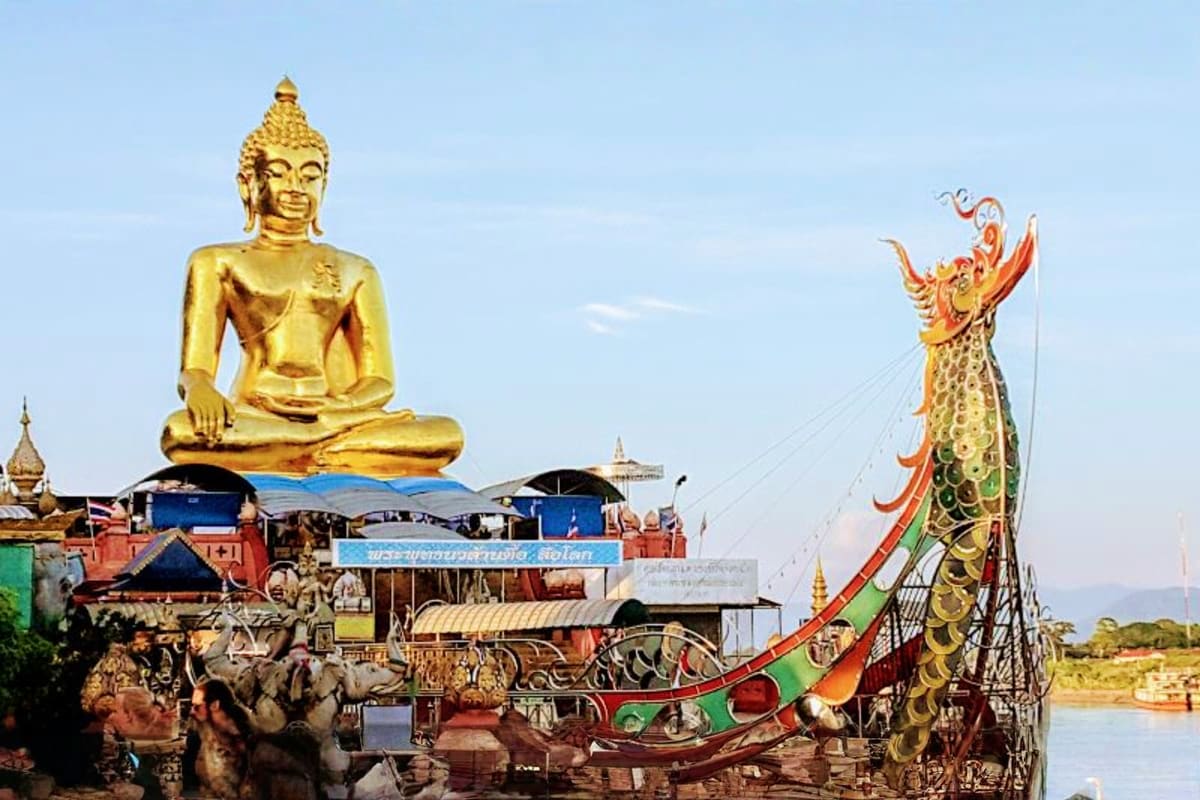 Golden Buddha statue seated in meditation pose with ornate dragon-headed boat on the Mekong River in Thailand
