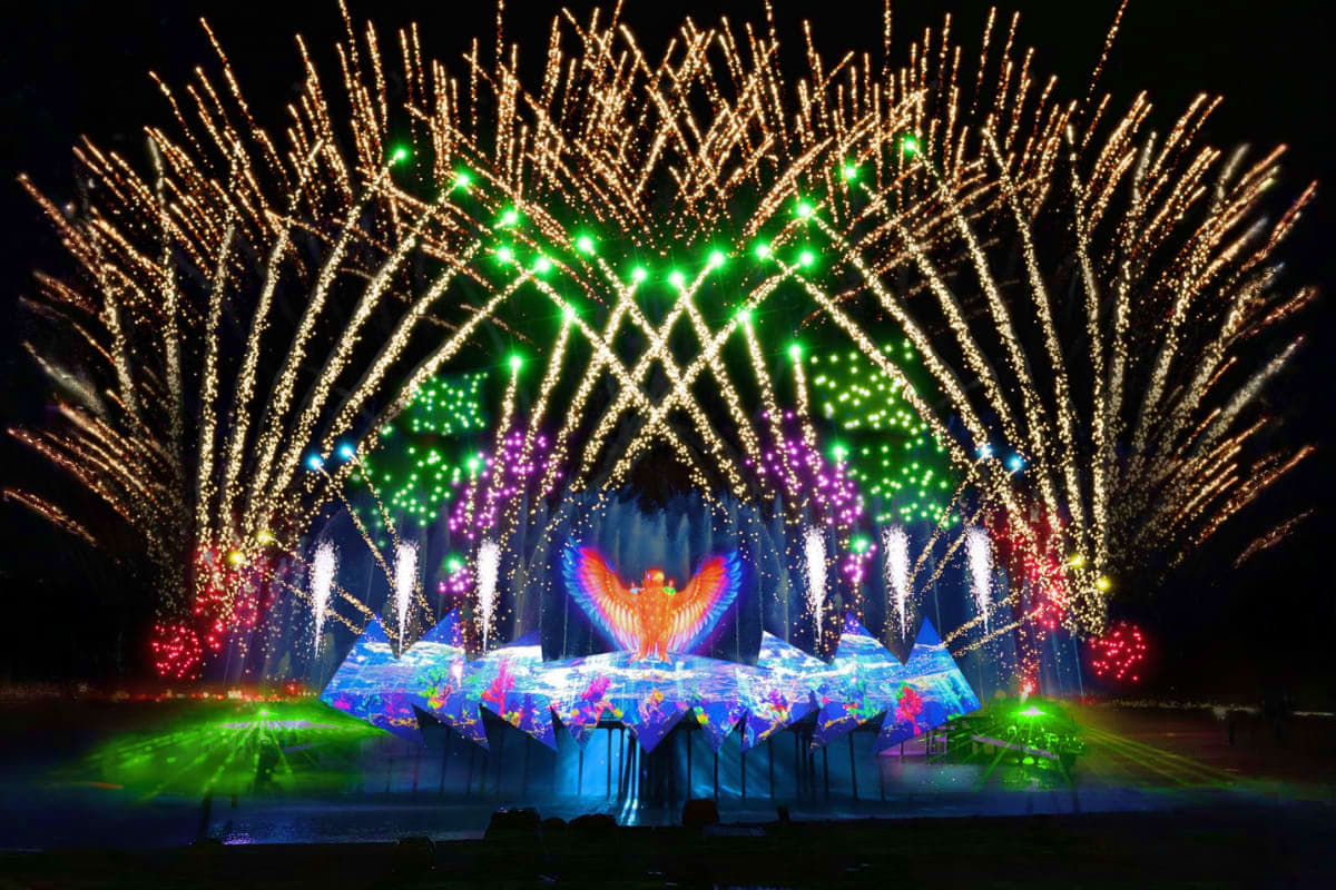 Elaborate nighttime light show stage display with illuminated phoenix figure, colorful LED panels, and arching golden light c