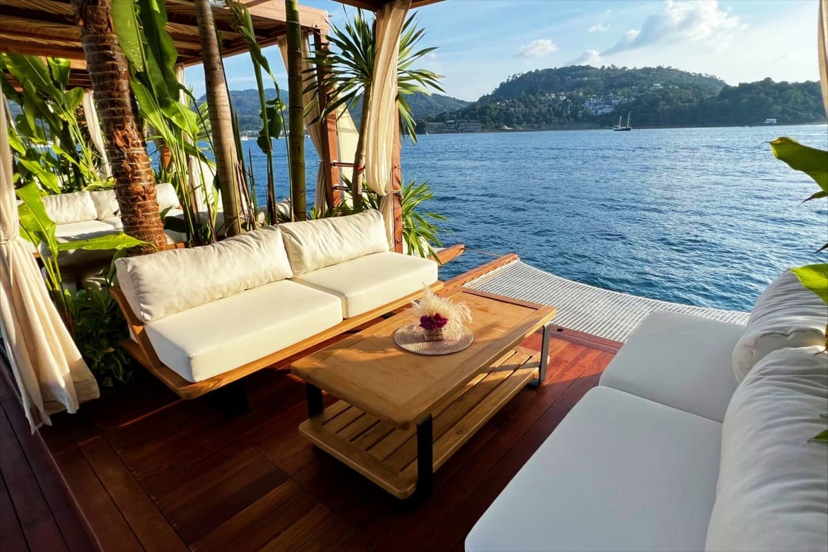 Yona Beach Club Luxury waterfront lounge with white cushioned seating, wooden deck, tropical plants, and bay views with forested hills in bac