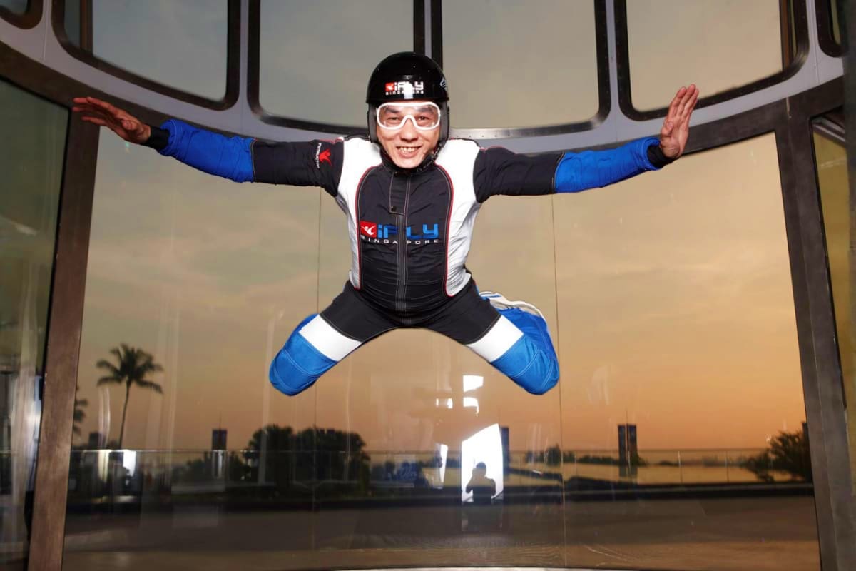 Person in blue and black flight suit mid-jump in indoor skydiving wind tunnel at iFly Singapore with scenic waterfront view v