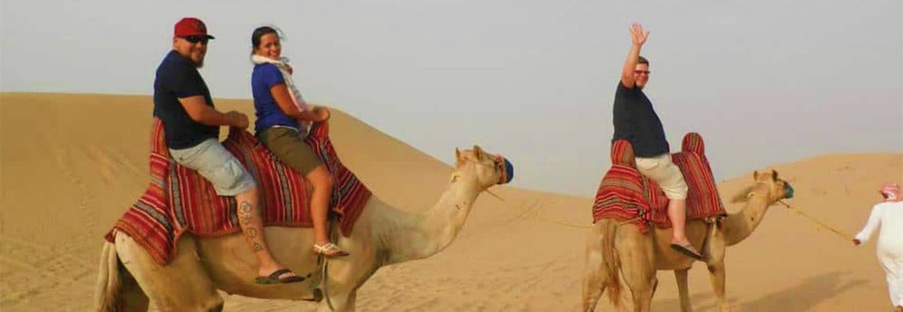 Jeddah Safari with Quad Bikes and Camel Ride Tourists riding camels in desert sand dunes near Jeddah, Saudi Arabia, wearing traditional red and white saddle blankets