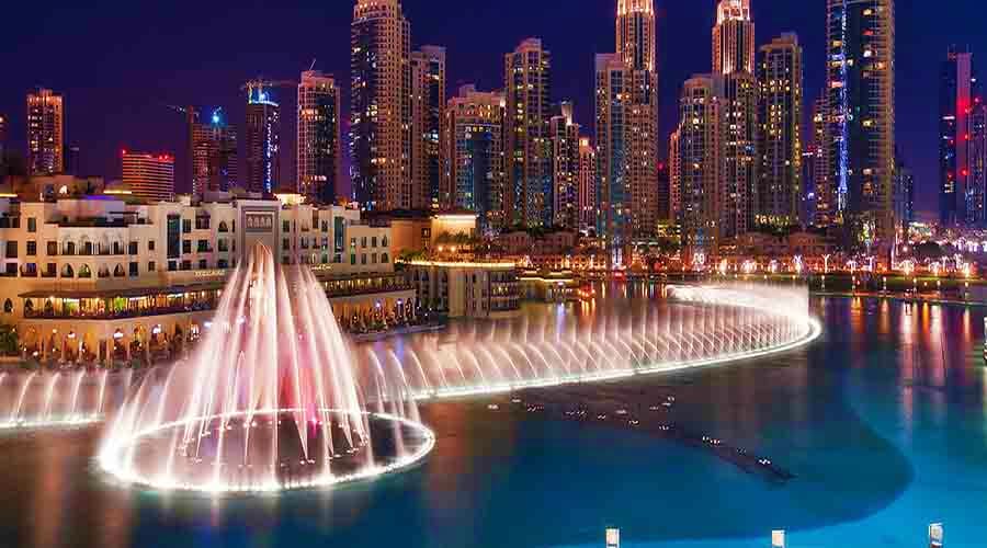 Illuminated Dubai Fountain performing at night with pink and white water jets, downtown skyscrapers reflected in the lake bel