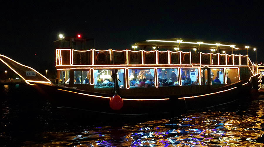 Illuminated traditional abra boat on Dubai Creek at night with passengers visible through windows, decorated with warm golden