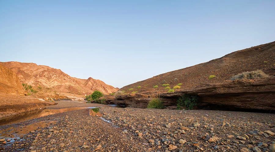Arid mountain landscape with rocky terrain, shallow water stream, and sparse desert vegetation in Hatta valley