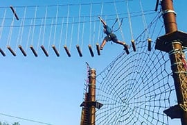 Person climbing rope obstacle course at adventure park with blue sky and wooden poles visible