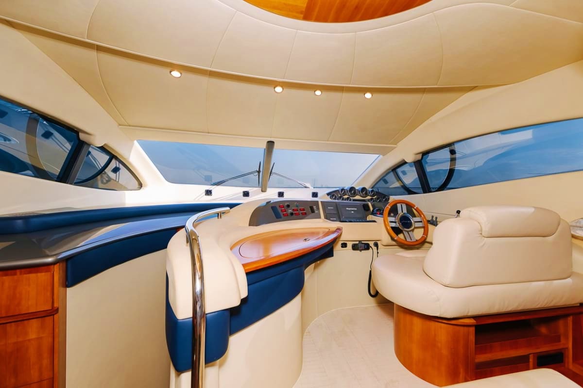 Luxury yacht cabin interior with cream upholstered seating, wooden accents, orange steering wheel, control panel, and large w
